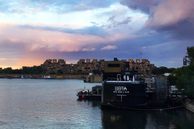 a lake with the Bota Bota structure floating on it and building in the background with a pink, purple and blue sky overhead