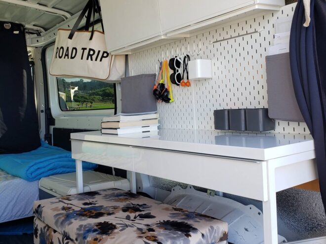 interior space of converted van, focusing on a desk area with ottoman and sunglasses hanging on the wall and a tote bag with the words "Road Trip" on it
