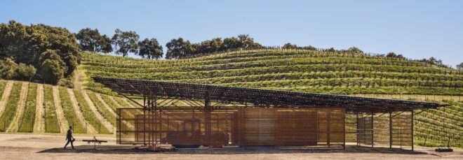 a landscape with a barn in the foreground, with a person walking towards it on the left. in the background are rolling hills with rows of greenery.
