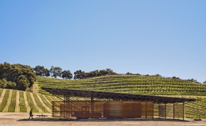 a landscape with a barn in the foreground, with a person walking towards it on the left. in the background are rolling hills with rows of greenery.