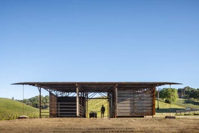 a landscape with a head-on view of a barn, with a person walking through the middle of it. in the background are rolling green hills.