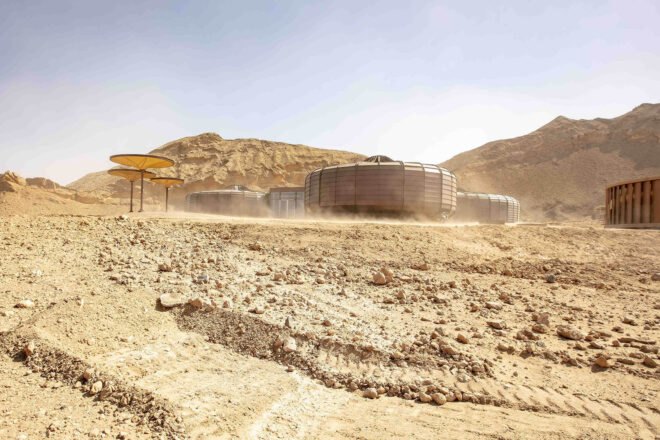 a ground angle of a desert landscape full of rocks with a dark brownish building composed of three structures, with three umbrella-like structures to the left and a small copper structure to the right