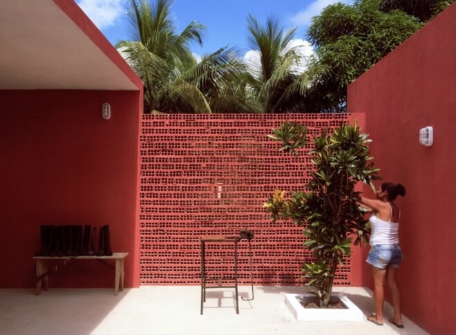 a red building with a fence-like structure linking two sections of building. to the left is a bench with several pairs of boots atop it. to the right is a green plant being pruned by a woman.