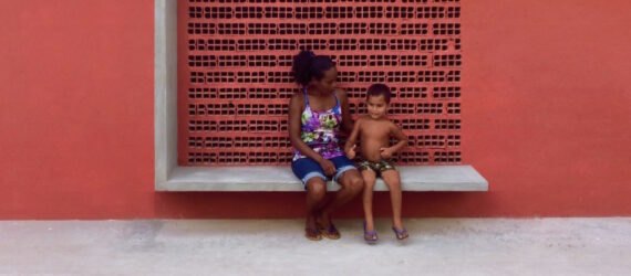 a red wall with a bench structure that a woman and child are citting on. a square cut-out behind them has a dotted pattern on the wall