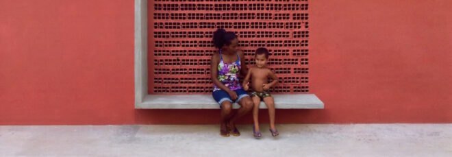 a red wall with a bench structure that a woman and child are citting on. a square cut-out behind them has a dotted pattern on the wall