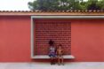 a red wall with a bench structure that a woman and child are citting on. a square cut-out behind them has a dotted pattern on the wall