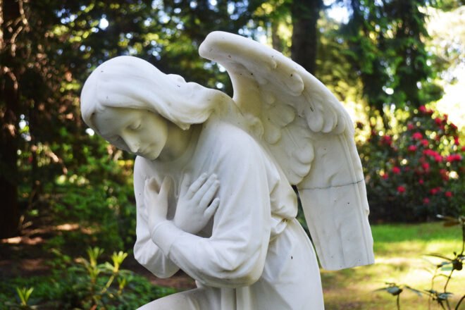 a white marble angel statue with its arms crossed over its chest. in the background is foliage.