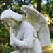 a white marble angel statue with its arms crossed over its chest. in the background is foliage.