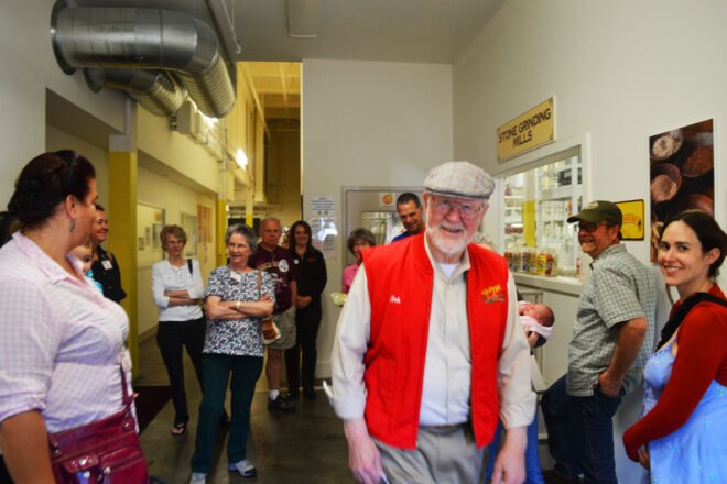 a group of people in a factory with an older person in flat cap and red vest looking at the camera