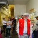 a group of people in a factory with an older person in flat cap and red vest looking at the camera