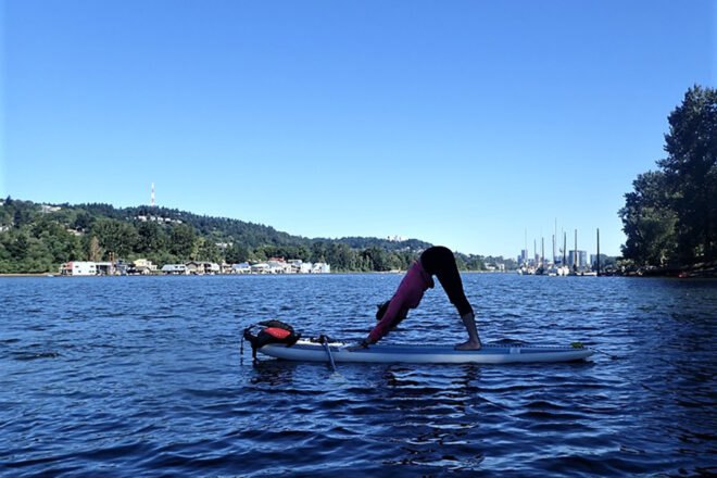 a lake with a person on a paddleboard who is doing yoga, in the background are green hills with houses on the coast