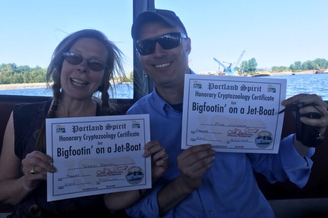 two people on a boat with the lake in the background. the two people are holding certificates that say "Portland Spirit, Honorary Cryptozoology Certificate for Bigfootin' on a Jet-Boat"