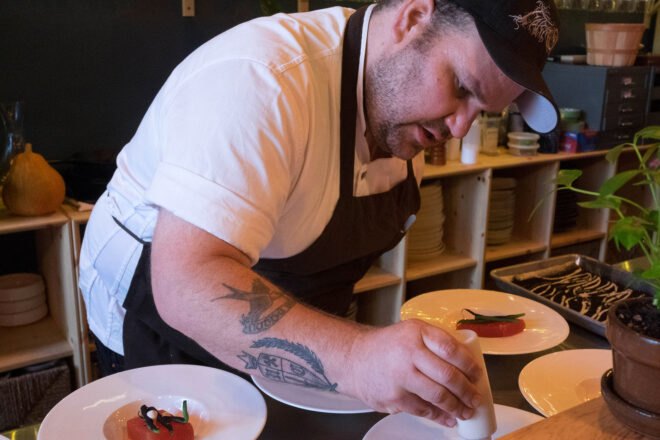 a person wearing a black cap and apron with white t-shirt. the person has tattoos on his arm, and he is holding a small bottle while he dresses plates of food