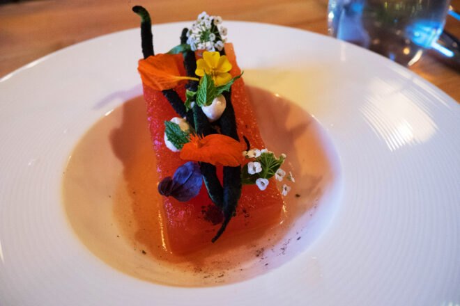an up-close shot of a plate of food, which features a rectangular piece of food that looks like watermelon, topped with three long black beans, flowers and leaves