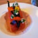an up-close shot of a plate of food, which features a rectangular piece of food that looks like watermelon, topped with three long black beans, flowers and leaves