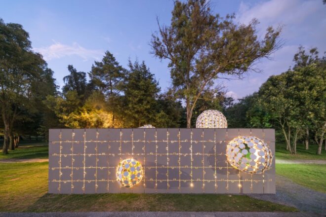 a night view of a short, gray rectangular building with a facade made of interlocking puzzle piece shapes. in the middle of the building is a protruding ball made up of several circular cut-outs. another, smaller ball protrudes from the right side, and another ball protrudes from the top left of the building. greenery surrounds the building. light from within the building filters through the open spaces in the facade