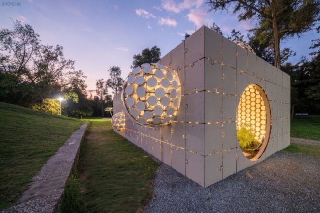 a side view of a short, gray rectangular building with a facade made of interlocking puzzle piece shapes. in the middle of the building is a protruding ball made up of several circular cut-outs. another, smaller ball protrudes from the right side, and another ball protrudes from the top left of the building. light from within the building filters through the open parts of the facade