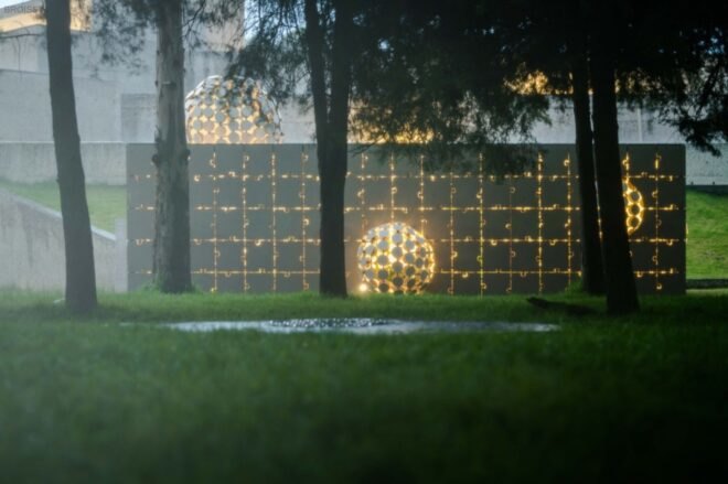 a night view of a short, gray rectangular building with a facade made of interlocking puzzle piece shapes. in the middle of the building is a protruding ball made up of several circular cut-outs. another, smaller ball protrudes from the right side, and another ball protrudes from the top left of the building. trees in front of the building partially obscure it. light from within the building filters through the open parts of the facade