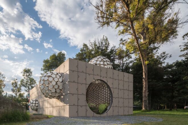 a side view of a short, gray rectangular building with a facade made of interlocking puzzle piece shapes. protruding balls made up of circular cut-outs dot the outside of the building. on one side of the building is a circular entrance that leads inside. surrounding the building is greenery and trees
