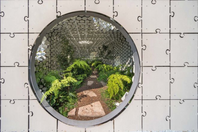 a close-up view of the circular entrance leading into the short gray building. inside the building are walls composed of circular cut-out shapes. also inside is a stone path through a lush, green plant-filled area.