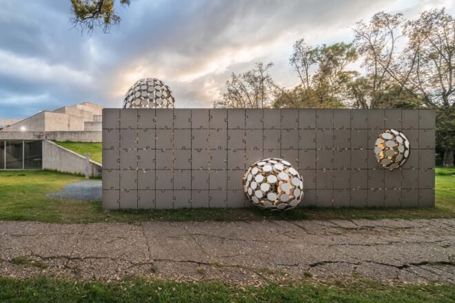 a short, gray rectangular building with a facade made of interlocking puzzle piece shapes. in the middle of the building is a protruding ball made up of several circular cut-outs. another, smaller ball protrudes from the right side, and another ball protrudes from the top left of the building. in the background are more buildings and greenery