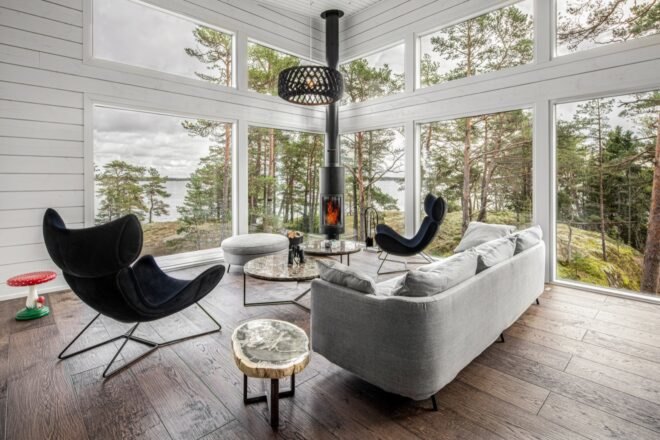 a living room with small fireplace, two chairs, tables and a sofa. large windows look out on trees and a body of water