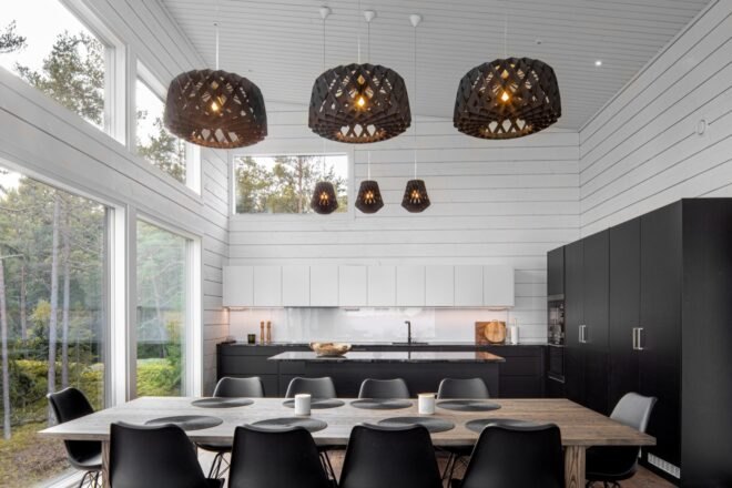a dining table with chairs, and three light fixtures above it. behind the table is a kitchen with black cabinetry to the right.