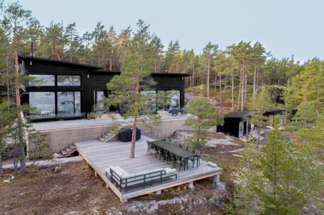 trees surround a house with a patio that has a tree growing out of the middle of it. a sofa and long dining table with chairs sits on the patio. two black shed-like structure are to the right. above the patio is a house with a black facade