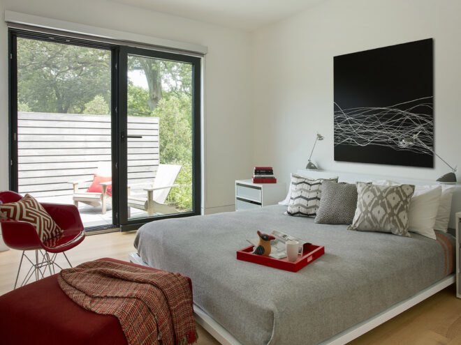 a bedroom with a red chair on the left and a gray bed on the right/ on the opposite wall is a sliding glass door