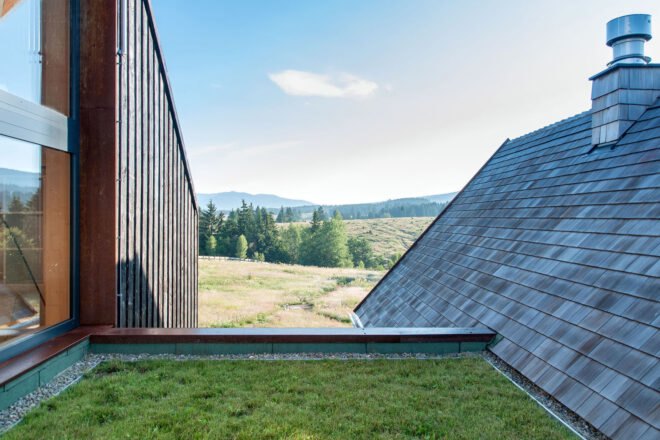 a green roof area in-between two parts of the building. in the background are rolling hills and green trees. to the right is a slate gray roof. to the right is a wood structure with windows.