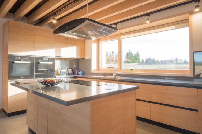 a kitchen full of light-colored wood cabinetry. a single, large light fixture hangs above an island in the middle of the kitchen. a bole os fruit sits on the countertop of the island. two ovens are built into the cabinetry. a large window near the sink looks out on the field outside.