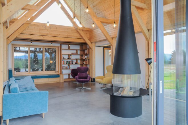 a living room with a modern fireplace that stands free in the room and rises up to the ceiling in a long black funnel-like structure. to the left is a blue sofa, in the background are purple and yellow chairs. above are wood, triangular structures that make up the ceiling.