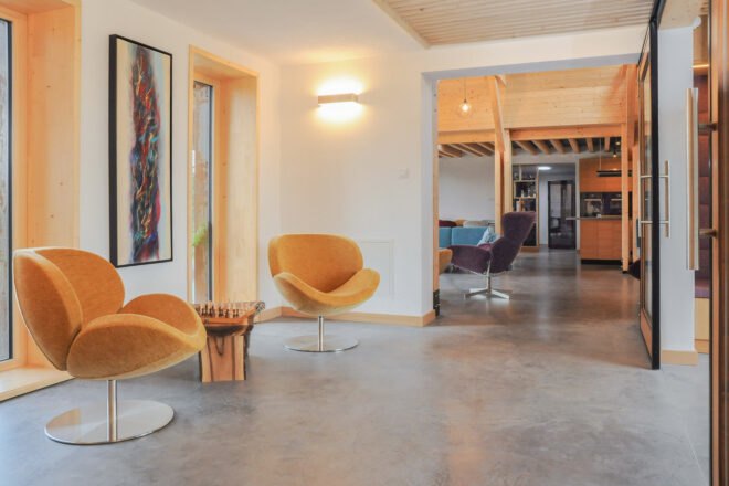 an interior shot of the house. to the left are two modern looking mustard yellow chairs, with a small wooden table between them and two windows behind the chairs on the wall. a long rectangular artwork is on the wall behind them. to the right you can see into another room with a kitchen and purple chair.