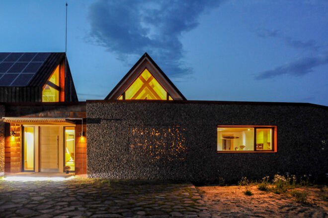 a night view of the outside of the house. a grey, textured facade is shown to the right of a brightly lit front entrance area. above you can see two windowed areas peaking out from structures behind the main structure, with lit up windows.