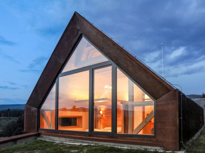 a triangular building with a rust colored frame around glass walls that look into the bedroom of the house.