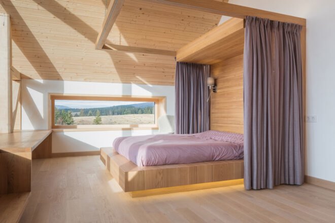 a bedroom with a purple bed on a raised platform with purple curtains surrounding it on either side. on the opposite wall is a horizontal rectangular window that shows a picturesque view of hills and trees.