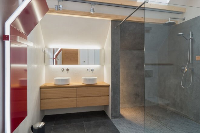 to the left, a red accent piece is built into the wall, forming a towel rack. a sink area in the middle of the room features wood cabinetry and a backlit mirror. to the left is a slate gray shower area with glass walls