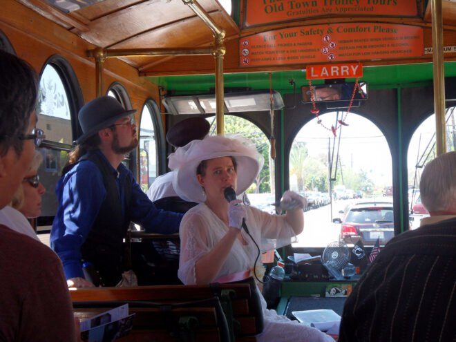 the interior of a trolley with two people guiding a tour at the front of the trolley. one tour guide wears a pink dress and hat, while the other person wears a suit and bowler hat