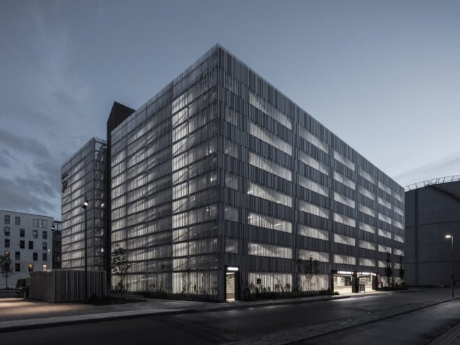 a night-time view of the back of a white-silver rectangular building. light filters through the slats on the building's facade