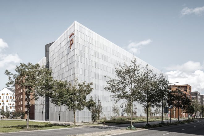a white-silver rectangular building with a P on it, marking it as a parking structure. the building is surrounded by trees and grass. a road is in the forgeround