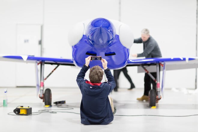 a person sits on the floor in front of a blue plane, taking a picture of it on a phone