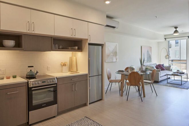 a kitchen with over, fridge and kettle on the stove. a dining table with four chairs is next to the kitchen and living room, where a sofa and lamp sit