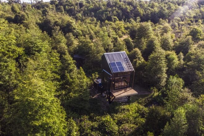 bright green trees surround a small, solitary cabin with a wood deck and solar panels on the roof