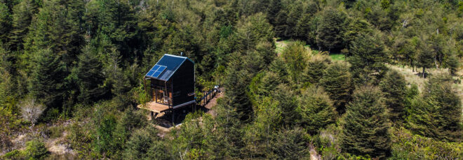 green trees surround a small, solitary cabin with a wood deck and solar panels on the roof