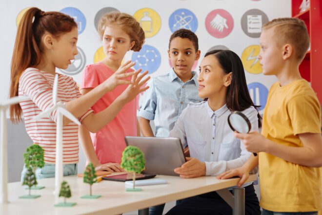 teacher listening to students talk with small trees and wind turbine models on a desk