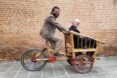 person in suit riding a bamboo bike with another person seated in the front