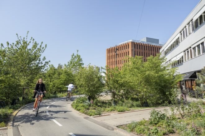 a green space with a bike path where people are riding bikes. a brown-orange building is in the background