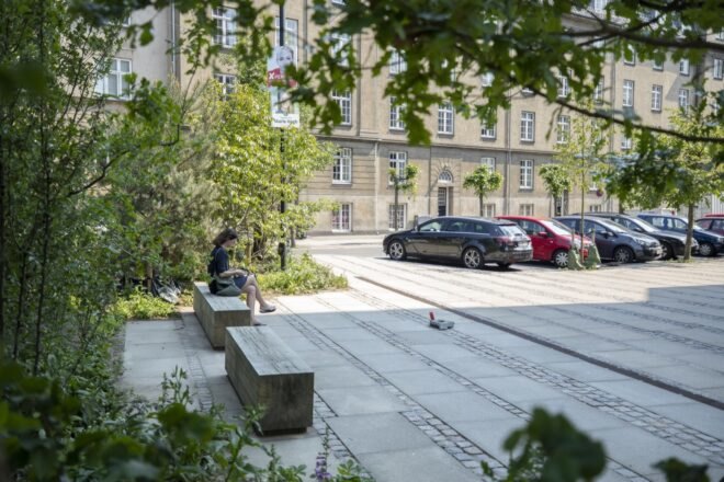 to the left a green space and benches with one person sitting. to the right is a parking area with cars.