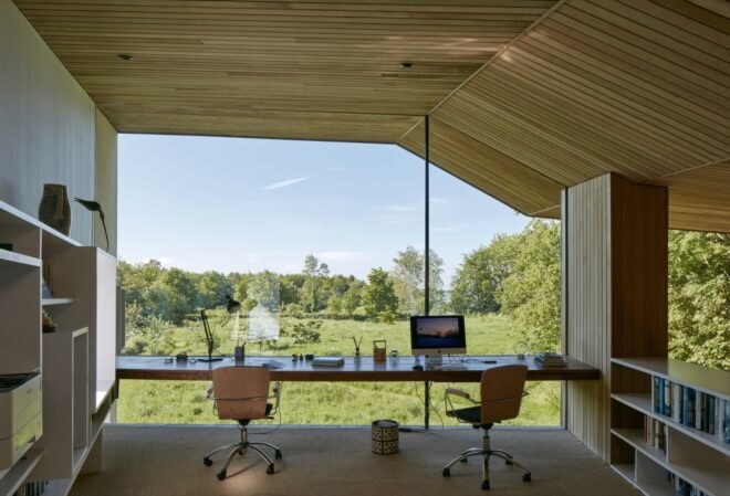 an office area with wood floor, walls and ceiling. a desk and two chairs look out through a floor-to-ceiling glass wall that shows greenery outside.