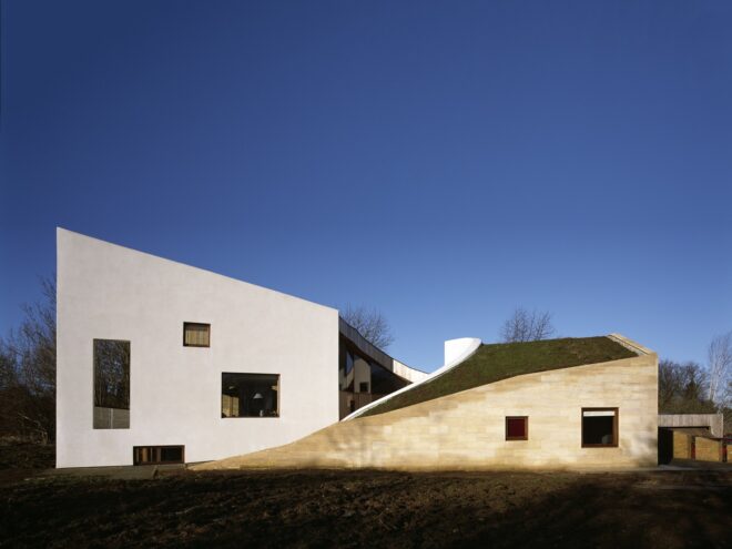 an angular building with a plain white facade on the left with three windows, and a sloped green roof visible on the right